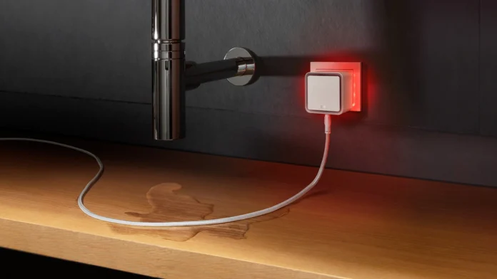 Een slimme WaterMelder in het stopcontact geeft een rood alarmsignaal wanneer de sensorkabel een plas water op de houten ondergrond detecteert.