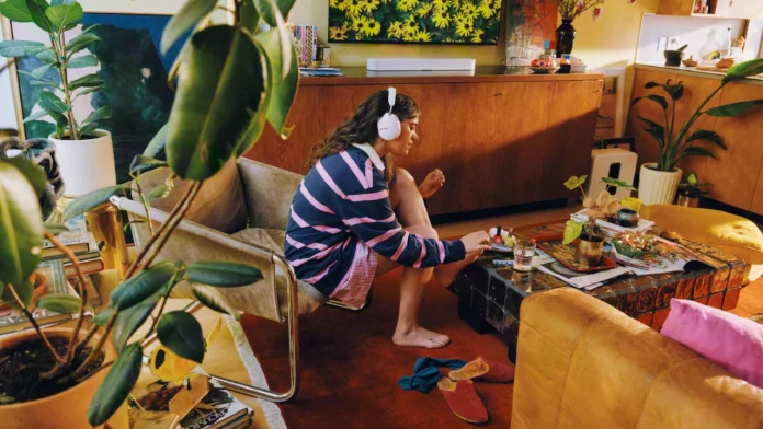 Woman wearing white Sonos headphones in a cozy living room with a Sonos soundbar and lush indoor plants.