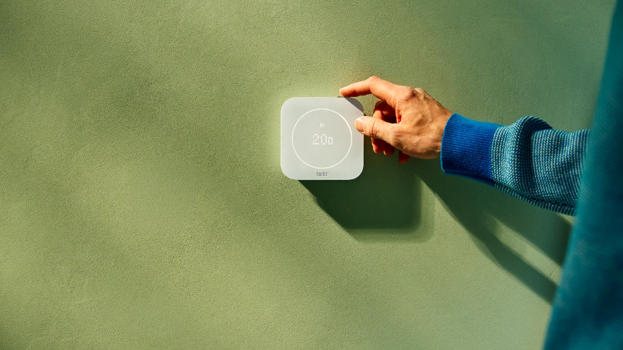 Een hand stelt de temperatuur in op een tado slimme thermostaat tegen een groene muur.