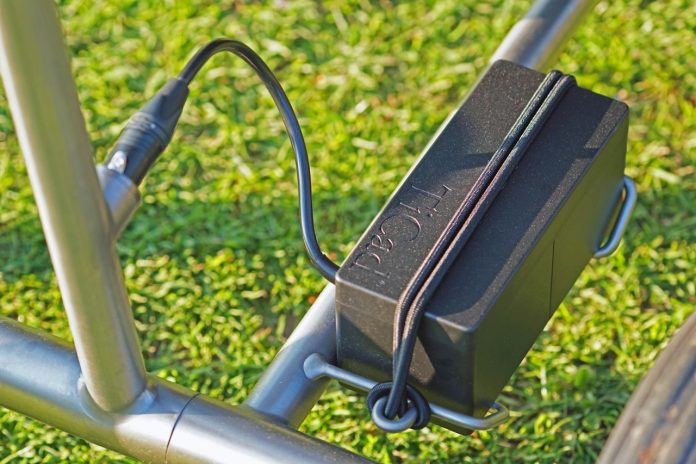 Close-up van een fietsaccu met laadkabel op het frame van een e-bike, met gras op de achtergrond.